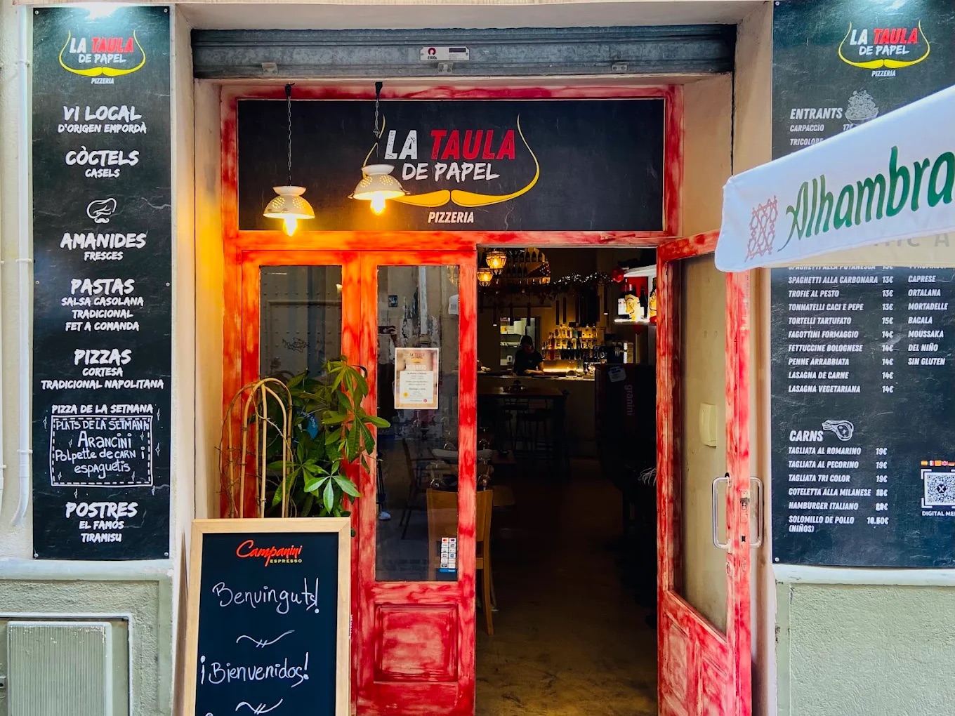 La entrada de La Taula de Papel con la puerta roja abierta: rótulo, pizarras con la carta y el interior al fondo